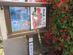 赤い生垣の前に設置された木製の掲示板に、マラソンを走る円谷幸吉氏の姿を描いた力強いイラストと、競技場の風景を鮮やかに描いた絵画が並んで展示されている、屋外の「物語ボックス」という案内板を斜めから捉えた写真