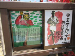 生垣の前に設置された掲示板に、手を振る人物のイラストと文、そして「東京オリンピックと円谷幸吉」と題されたランナーの力強い絵が並んで展示されている、物語ボックス」の写真