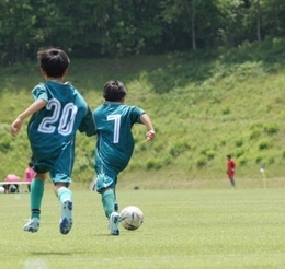 緑の芝生の上で、サッカーボールを追いかけて走る緑色のユニフォームとソックスを身に着けた男の子2人の後ろ姿の写真