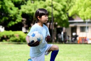 屋外の芝生の広場で、水色のユニフォームを着てサッカーボールを片手に抱え、片膝をついている子どもの写真