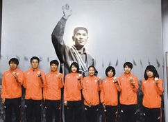 オレンジ色のジャージを着た8人が横一列に並び、全員が右手で軽く拳を握ってポーズを取り、背景には右手を挙げてメダルをかけた円谷幸吉の大型白黒写真が壁一面に飾られている室内の写真