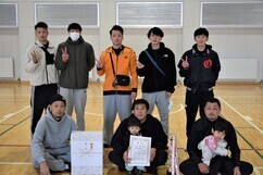 体育館のコートにて、賞状を掲げる男性や小さな子供を抱いたメンバーなど、カジュアルな服装の男性たちが二列に並び、手にしたトロフィーや大きな白い副賞の箱を囲んで穏やかな表情で撮影に応じている大会の記念写真