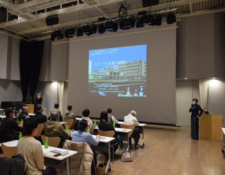 広いホールに並んだ机と椅子にたくさんの参加者たちが座る中、前方の大きなスクリーンに資料が映し出され、右横の演台前にマイクを持った講師の女性が立ち、話をしている様子の写真