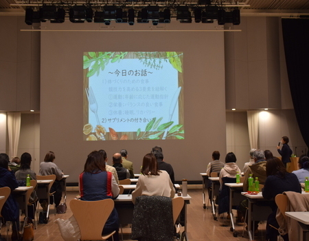 会場に並んだ2人掛けのテーブルに着席した参加者たちが、前方のスクリーンに映し出された「今日のお話」と題された資料を見ている様子を後方から撮影した写真
