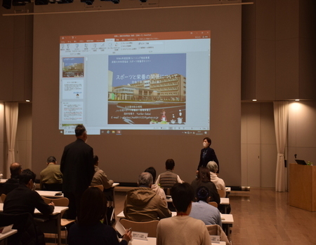 資料が映し出された大きなスクリーンの前に、講師の女性が立ち、参加者席から立ち上がった男性が講師に質問をしている様子の写真