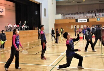 体育館で大勢の人々がコートに立っており、手前では紺色とピンクの配色のユニフォームを着た数人の男女がファミリーバドミントンを行っている様子の写真