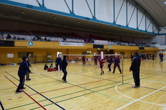 広い体育館に設置されたコートで、ラケットを手に構える選手たち3名がネット越しに向かい合い、高く上がったシャトルを見上げながらプレーに集中している、バドミントン大会の試合風景を捉えた写真