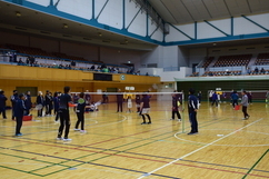 広い体育館に設置された複数のコートで、多くの選手たちがラケットを手にシャトルを追い、ネット越しに白熱した試合を繰り広げているバドミントン大会の全体風景を捉えた写真