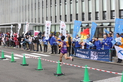 沿道に並ぶ多くの観客や青いジャンパーを着たスタッフ、火の形をしたマスコットキャラクターに見守られながら、紫色のユニフォームを着たランナーが道路を力強く駆け抜けていく駅伝大会の風景の写真