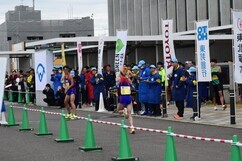 沿道に立ち並ぶスポンサー企業ののぼり旗の前を、青いウェアを着た大会スタッフや観客たちが見守る中、ゼッケンをつけたランナーが力走し次のランナーに襷を渡す中継所の場面を捉えた写真