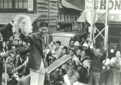 首にメダルをかけ、大きな花束を持った円谷幸吉選手がオープンカーの座席に立って集まった観衆に手を振っている凱旋パレードの写真