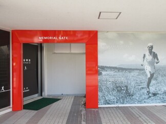 畦道を走っている円谷幸吉選手の大きな白黒写真が一面に貼られた壁の横に赤い枠で囲まれた入口ゲートがある写真