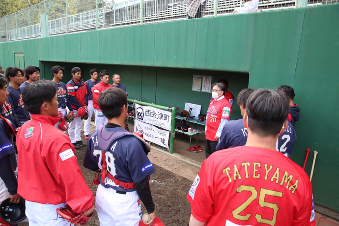 濃紺や赤のユニフォーム姿の野球選手たちが、背番号30の赤いユニフォームを着た安藤副社長を囲み、ベンチ前で話を聞いている様子