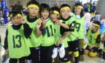 試合会場を背景に、蛍光グリーンのビブスをつけ、黄色の鉢巻きを巻いた6人の子どもたちがカメラに向かってピースサインをしている写真