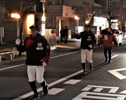 日が落ちた街中の道路を右手に松明を持った野球のユニフォームを着た男性たち3人が走っている様子の写真