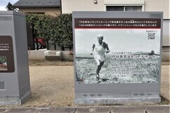 草原を走る幸吉選手のモノクロ写真とともに説明書きが記された、屋外に設置された四角いグレーの配電用機器の写真