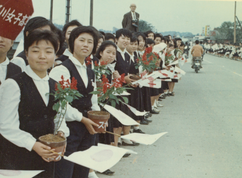 沿道に並んだ制服姿の女子学生たちが、赤いサルビアの花の鉢植えと日の丸の旗を手に持ち、カメラの方を向いている写真