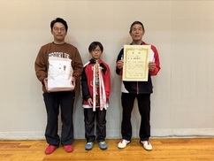 バドミントン大会の表彰式にて、賞品を抱えた茶色のスウェットの男性、中央でトロフィーを持つ子供、賞状を掲げる赤色のジャージを着た年配の男性の三世代と思われる三人が、体育館の壁を背に一列に並んで立つ記念写真