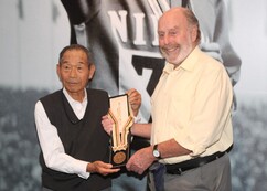 二人の人物が並んで立ち、中央で長方形の木製盾に取り付けられたメダルを共同で持っている様子が写され、背景にはスポーツジャージ姿の人物が大きく印刷された白黒写真が飾られている室内の写真