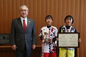 茶色の壁の前で、橋本市長と、ユニフォームを着てメダルを首にかけ、トロフィーと賞状を持った大武姫菜さん・夢菜さん姉妹が並んだ記念写真