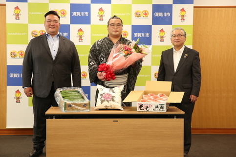須賀川市のロゴが並ぶバックパネルの前で、花束を抱えた力士の白熊関を中心に、スーツ姿の二所ノ関親方と橋本市長が並び、手前のテーブルには特産品のきゅうりや桃、米袋が贈呈品として並べられた、表敬訪問時の記念写真
