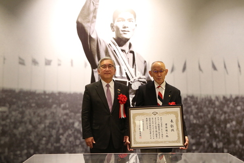首にメダルをかけた円谷幸吉選手のモノクロ写真のパネル前で、表彰状を持った君原さんと橋本市長が並んで立っている記念写真