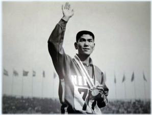 競技用ジャージを着た円谷幸吉選手が胸にメダルをかけ、右手を高く挙げている姿が写され、背景には多数の旗が並ぶ屋外の競技場が広がる白黒写真