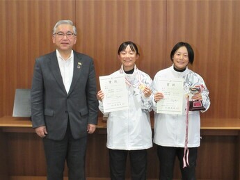 茶色の壁の前で、橋本市長と、白い上着を着て、メダルやトロフィーと賞状を持った大武姫菜さん・夢菜さん姉妹が並んだ記念写真