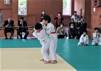 緑とベージュの配色のマットが敷かれた会場で、関係者が周りで見守る中、柔道着を着た男の子2人が試合を行っている様子の写真
