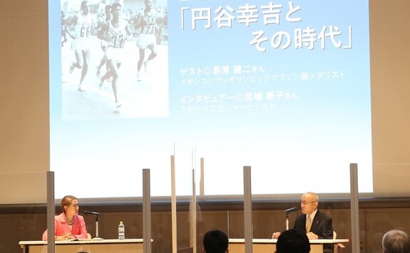 複数のアクリル板が設置されたトークイベント会場の壇上で席に座った男女が対談をしており、背後の大きなスクリーンには「円谷幸吉とその時代」と題された資料が映し出されている写真