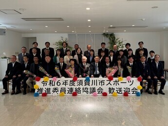 「令和6年度須賀川市スポーツ推進委員連絡協議会歓送迎会」の看板を前に、スーツ姿の男女が三列に並んで座り、前列の数名が贈呈された花束を大切そうに抱えながら穏やかな表情で記念撮影に応じている集合写真