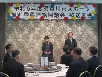 「令和6年度須賀川市スポーツ推進委員連絡協議会歓送迎会」の横断幕が掲げられた会場で、円卓を囲む出席者たちが見守る中、正面のマイクの前に立った女性が会合の挨拶やスピーチを行っている様子を捉えた写真