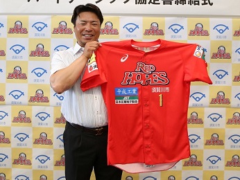 黄色と白のバックパネルの前に立ち、笑顔で須賀川市のロゴがついた赤いユニフォームを持つ岩村監督の写真