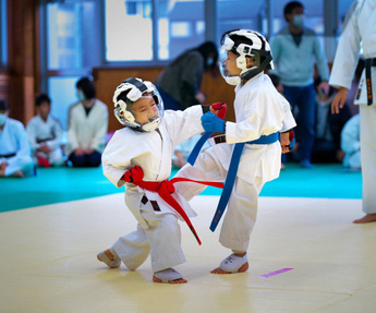 ヘッドガードをつけた、空手着に赤い帯を巻いた小さな子どもと、青い帯の子どもが空手の試合を行っている様子の写真