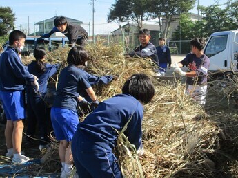 トラックの荷台いっぱいに積まれた枯れた萱を選手らと子どもたちが一緒に降ろしている様子の写真