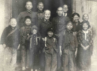 建物の前で男性たちと、着物を着た女性、7人の笑顔の子供たちが集まり、肩を寄せ合って写したモノクロの記念写真