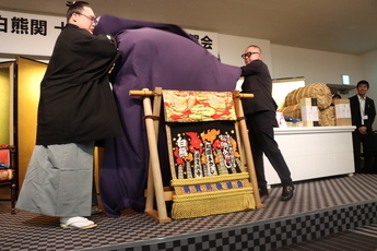 「白熊関十両優勝・新入幕祝賀会」のステージ上で、紋付き袴姿の白熊関とスーツ姿の男性が、木製の枠に掛けられた新しい化粧まわしを覆っていた紫色の布を左右から一気に引き上げ、華やかにお披露目している瞬間を捉えた写真