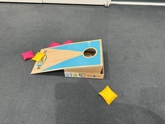 グレーのカーペットの上に、水色と薄い茶色の木目のカラーのコーンホールボードが斜めに置かれており、その周りに鮮やかな黄色とピンク色の小さな四角い袋が散らばっている写真