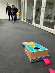 グレーのカーペット敷きの室内で、二人の男性がコーンホールゲームをしており、手前のコーンホールボードの穴に向かって、離れた場所から一人の男性が黄色い袋を投げている様子の写真