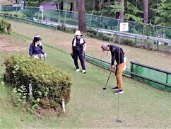 「7」と書かれた旗が立つパークゴルフ場のグリーンにて、茶色のパンツを履いた男性が慎重にパッティングを行っており、白い帽子を被った女性を含む二人の同行者がその後方で見守っている、木々に囲まれた屋外スポーツ風景の写真