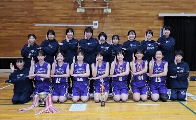 紫色のユニフォームや紺色のジャージ姿の女子メンバーが優勝カップやトロフィーを並べた後ろに前後2列に整列し、ウルトラマンのポーズをしている写真