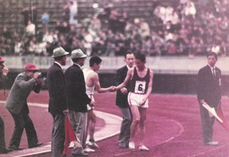 レース後の陸上競技場のトラックの上で、周囲には審判員やカメラマン、そしてスタンドを埋め尽くす大勢の観客が見守っている中で、背番号6をつけた君原選手と白のウェアを着た円谷選手が握手を交わしている写真