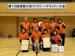 「第15回須賀川市ファミリーバドミントン大会」の看板の下で、お揃いのオレンジ色のTシャツを着た男女9名のチームが、優勝トロフィーと2枚の賞状を手に体育館のコート上で笑顔で並んでいる記念写真