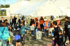 屋外の特設テントに「給水」と書かれた札が掲げられ、オレンジ色のベストを着たボランティアスタッフたちがランナーのためにコップに水を用意し、周囲には走り終えた参加者たちが集まっているマラソン大会の給水所の風景写真