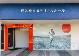 「円谷幸吉メモリアルホール」と書かれた青い看板の下に、ランニング姿の円谷選手の大きなモノクロ写真が壁一面に展示され、その左側にオレンジ色の枠で囲まれた「MEMORIAL GATE」という入口がある施設の外観の写真