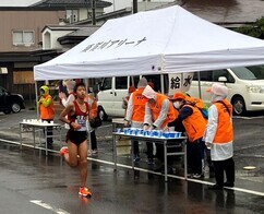 雨が降るアスファルトの道路上にて、えんじ色のユニフォームを着たランナーが、「給水」と書かれた白いテントの横を通り過ぎ、オレンジ色のベストを着たスタッフたちが紙コップを用意して待ち構えているマラソン大会の風景の写真