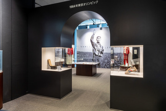 「1964年東京オリンピック」と書かれた黒い壁のアーチ越しに、当時の選手の巨大な写真や、ショーケース内に展示されたユニフォーム、競技用の靴、椅子などが並ぶ博物館の展示風景を捉えたカラー写真