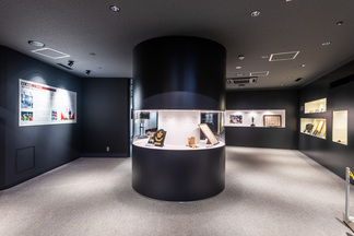 黒を基調とした展示室の中央に円柱型のショーケースが配置され、壁面には解説パネルや複数の埋め込み式展示スペースが並び、モダンで落ち着いた雰囲気の中で歴史的な資料を鑑賞できる博物館の内観写真