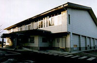 夕方の柔らかな光に照らされた、白い外壁と大きな切妻屋根を持つ二階建ての建物で、一階部分には等間隔に柱が並び、左端にある入口には雪よけのような小さな屋根とスロープが設置されている、静かな佇まいの公共施設の外観の写真