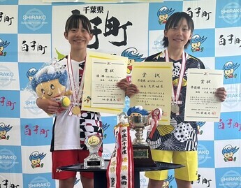 千葉県白子町のバックボードの前に、首にメダルをかけ、複数の賞状を持ちマスコットキャラクターのぬいぐるみを持ったユニフォーム姿の大武夢菜さん、姫菜さんが並んで立ち、手前に置いた大小2つのトロフィーが並んだ台越しに撮影した記念写真